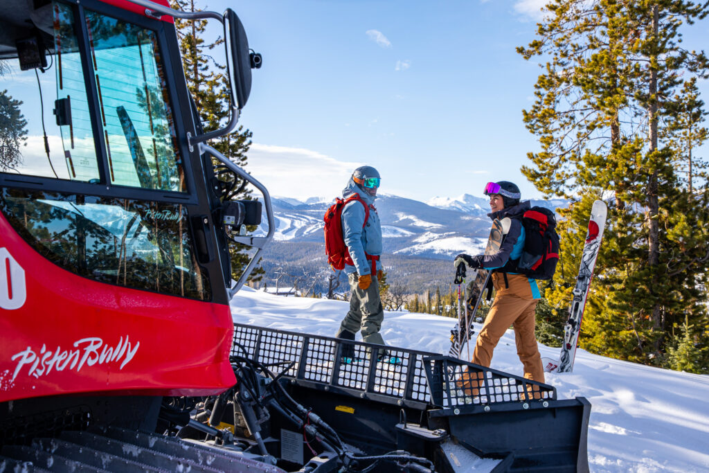 Two skiers stand by a snowcat with mountain and forest views.