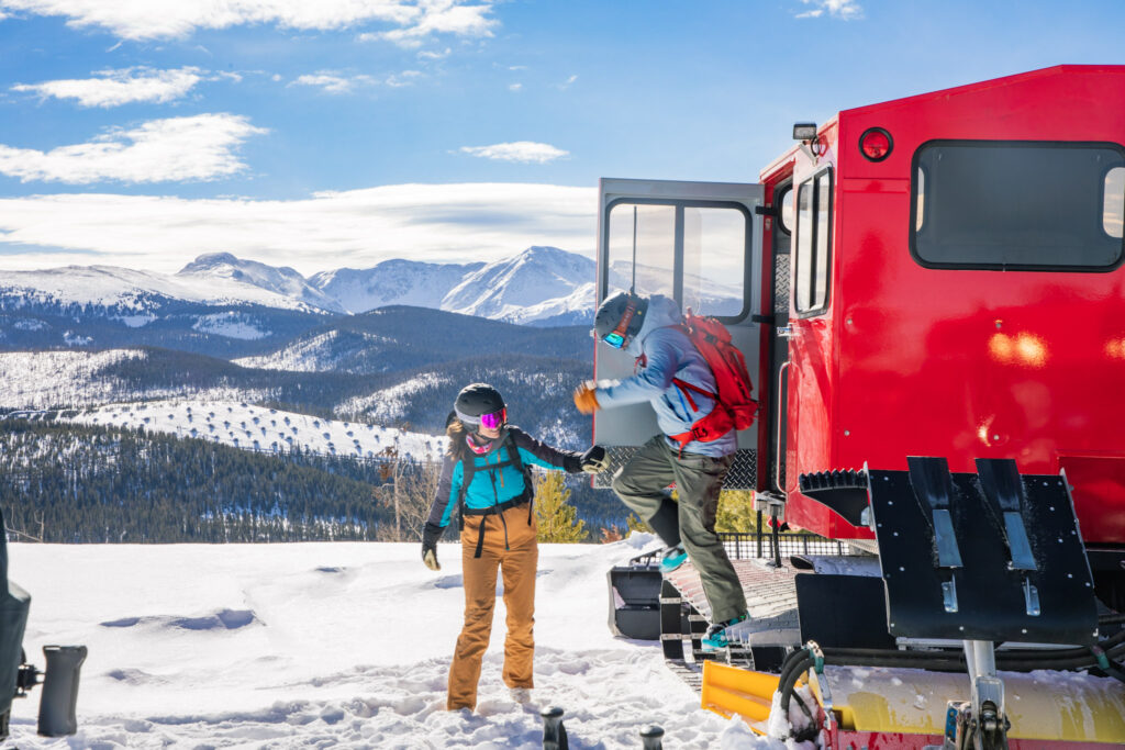 Two skiers exit a snowcat surrounded by mountain views.