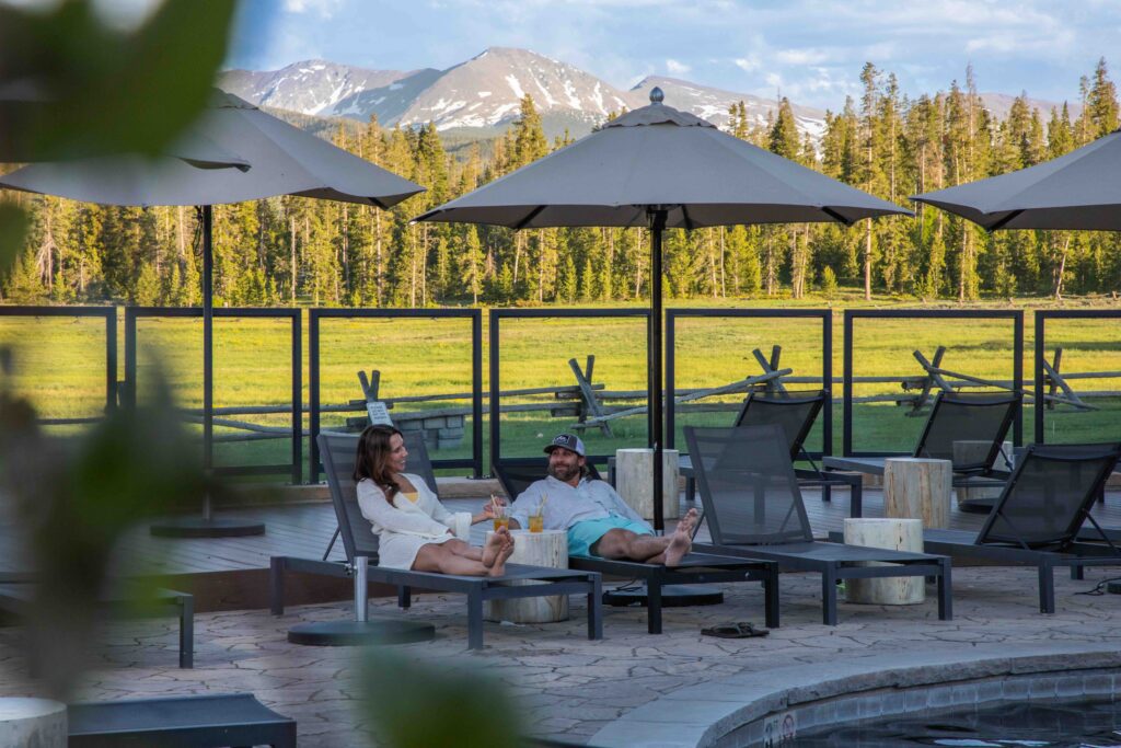 Two people lounge by the pool with drinks and mountains in the background