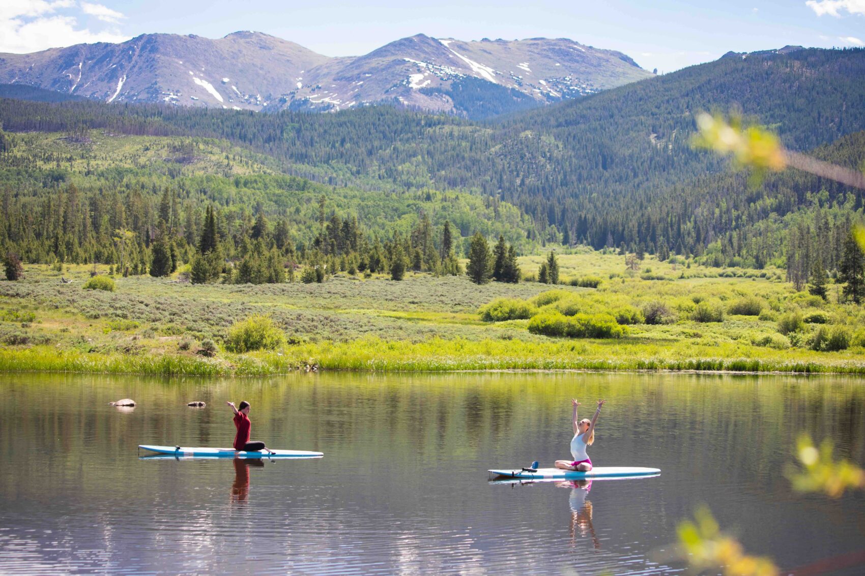 Two people stretch while floating on stand up paddle boards on a mountain pond with mountains in the background