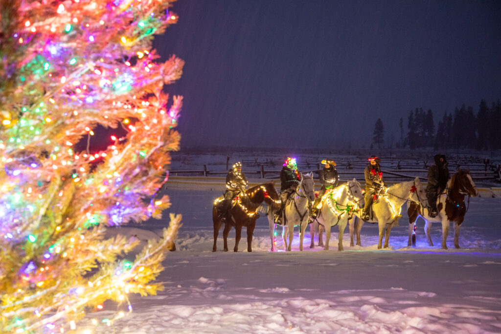 Outdoor holiday celebration with lit Christmas tree and riders on horseback with holiday lights.