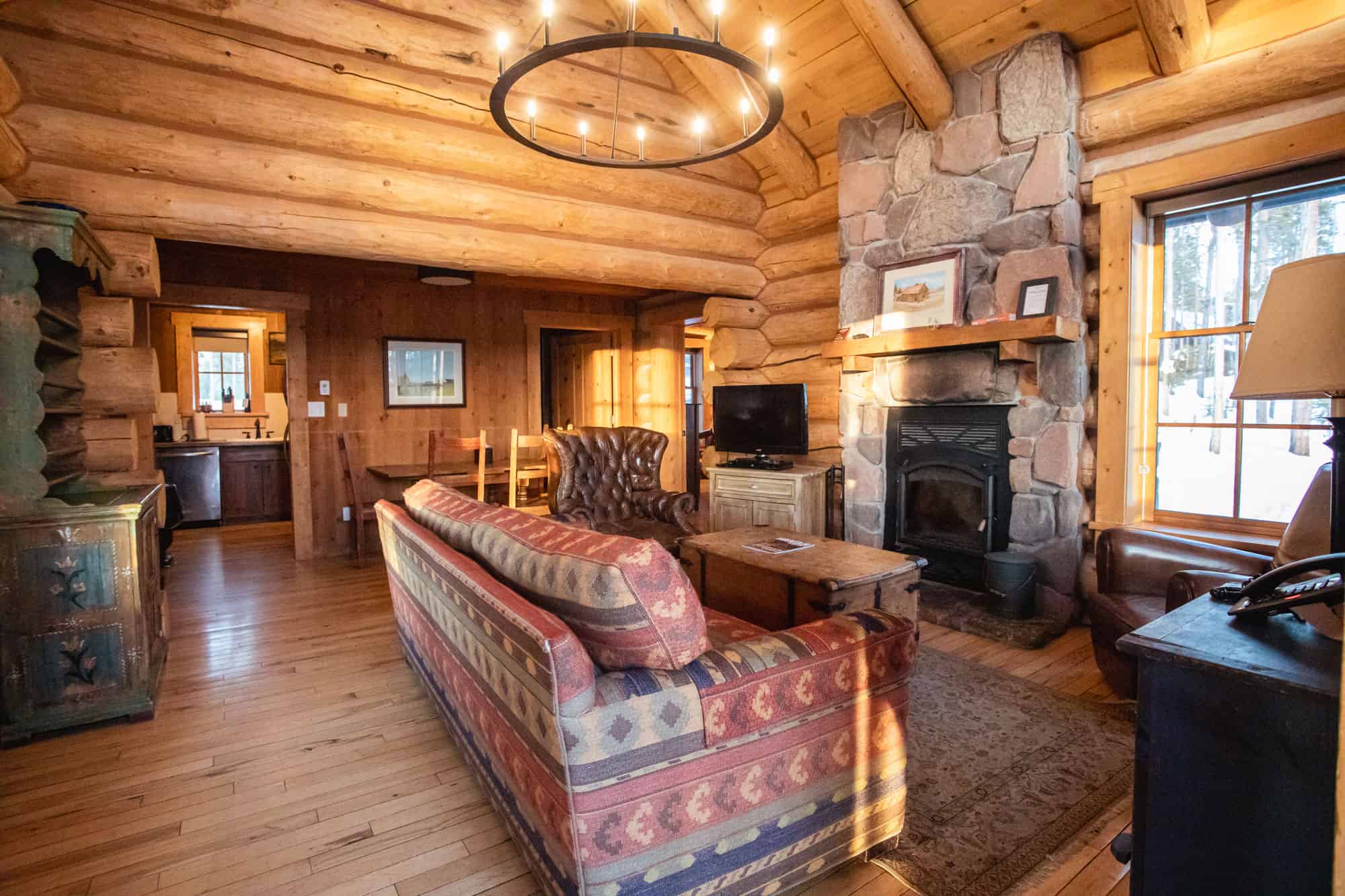 Warm log cabin living room with stone fireplace, wooden beams, and rustic furniture.