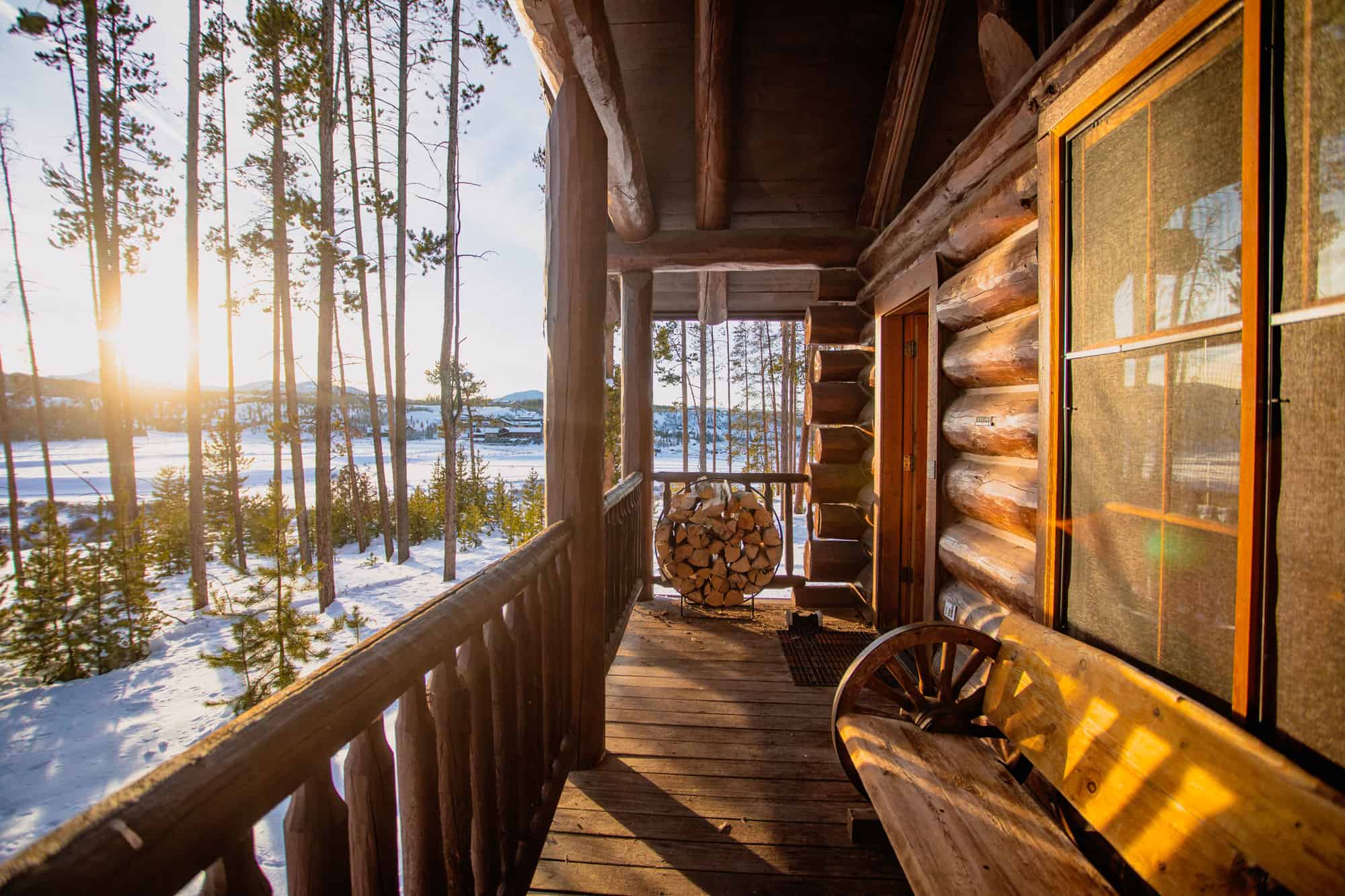 Rustic cabin porch overlooking snowy forest at sunset, with wood logs and cozy seating.