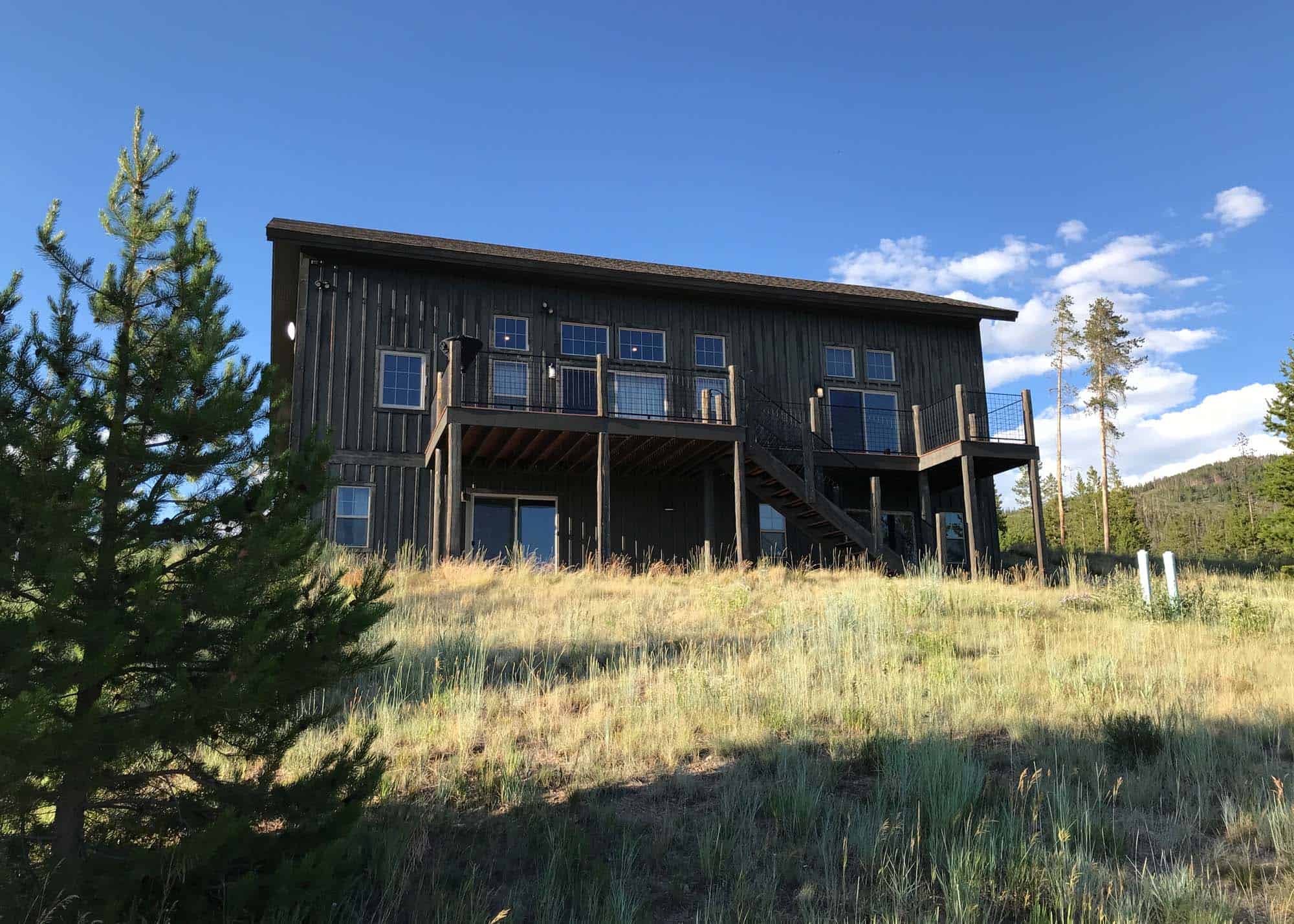 Modern black cabin with large deck on grassy hillside under blue sky.