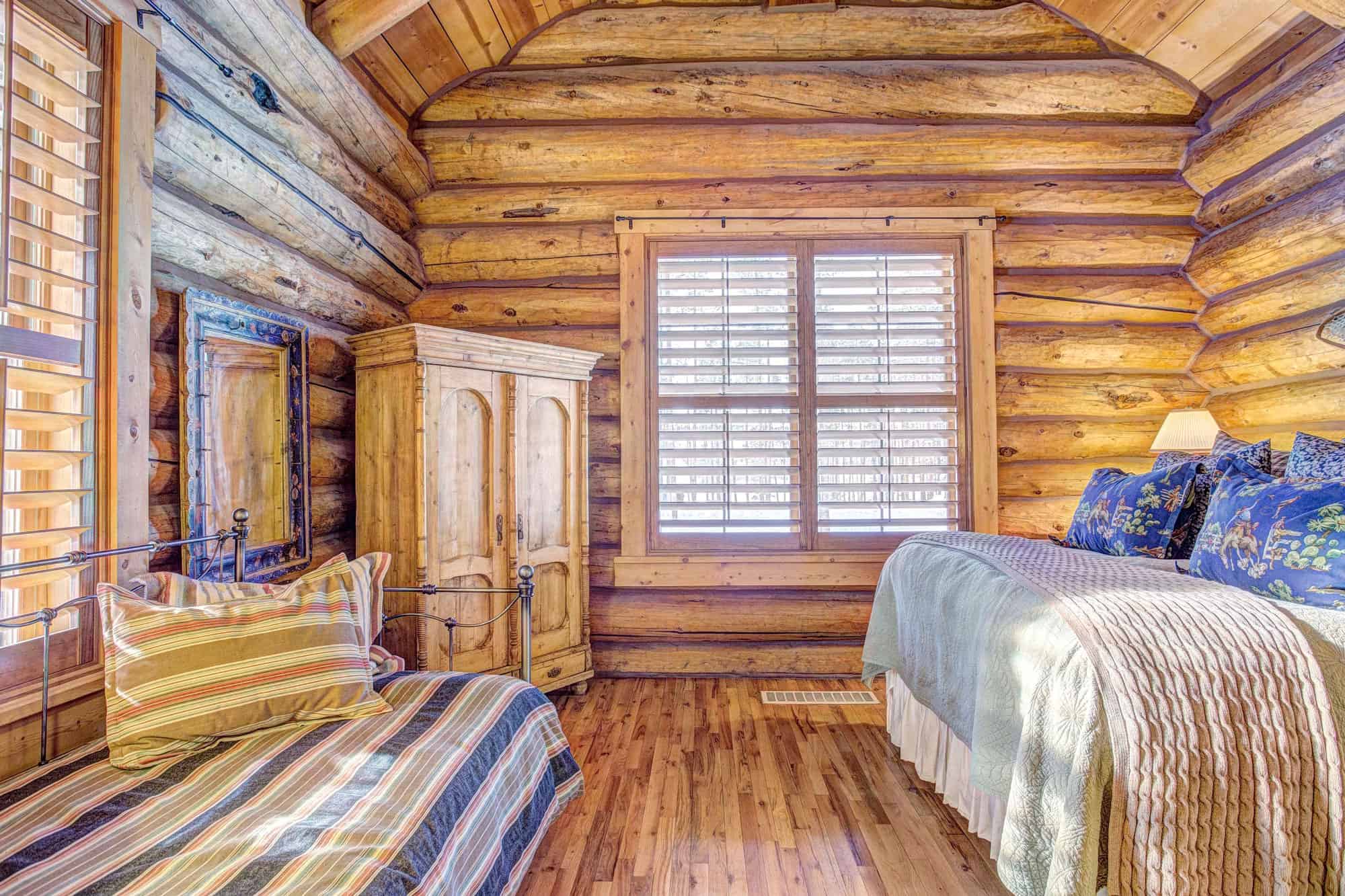 Charming rustic bedroom featuring wooden log walls, a large window with shutters, and comfortable be.