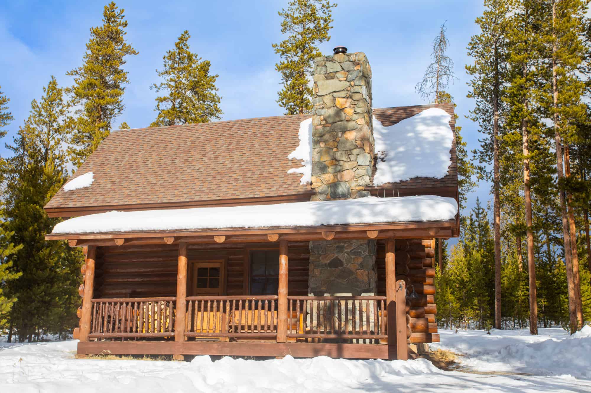 Cozy mountain cabin with snow-covered roof and stone chimney in a forest setting.