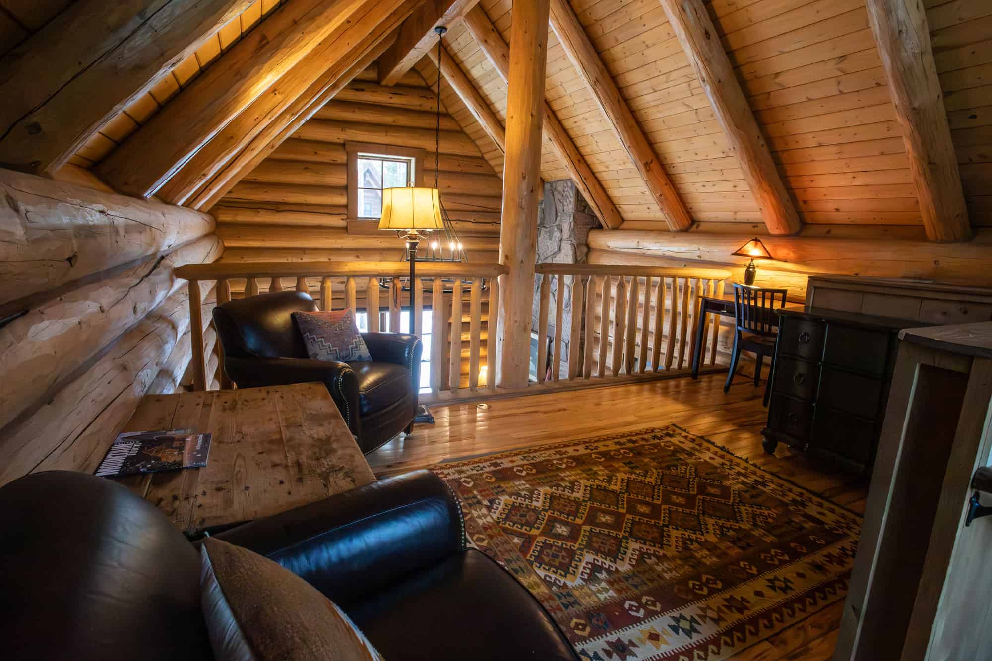 Interior view of a rustic log cabin loft with leather chairs, wooden beams, and cozy lighting.