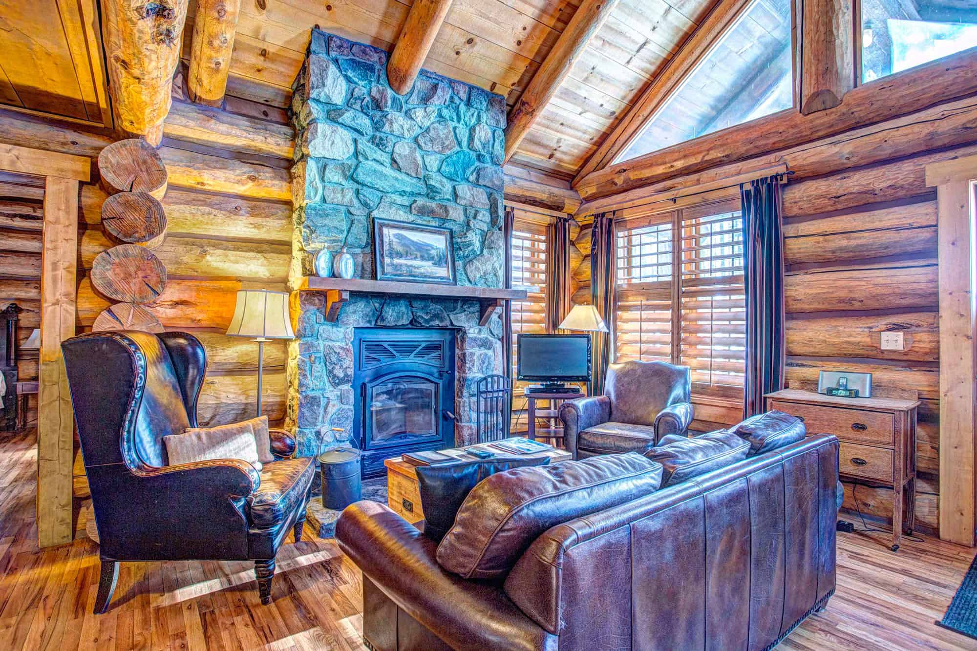 Warm log cabin living room with stone fireplace, leather sofas, and wooden accents.