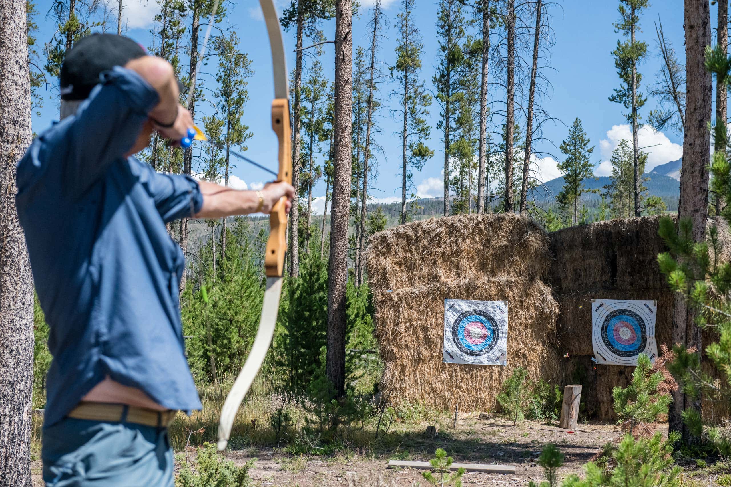 Person aiming a bow at outdoor archery targets in a forest setting.