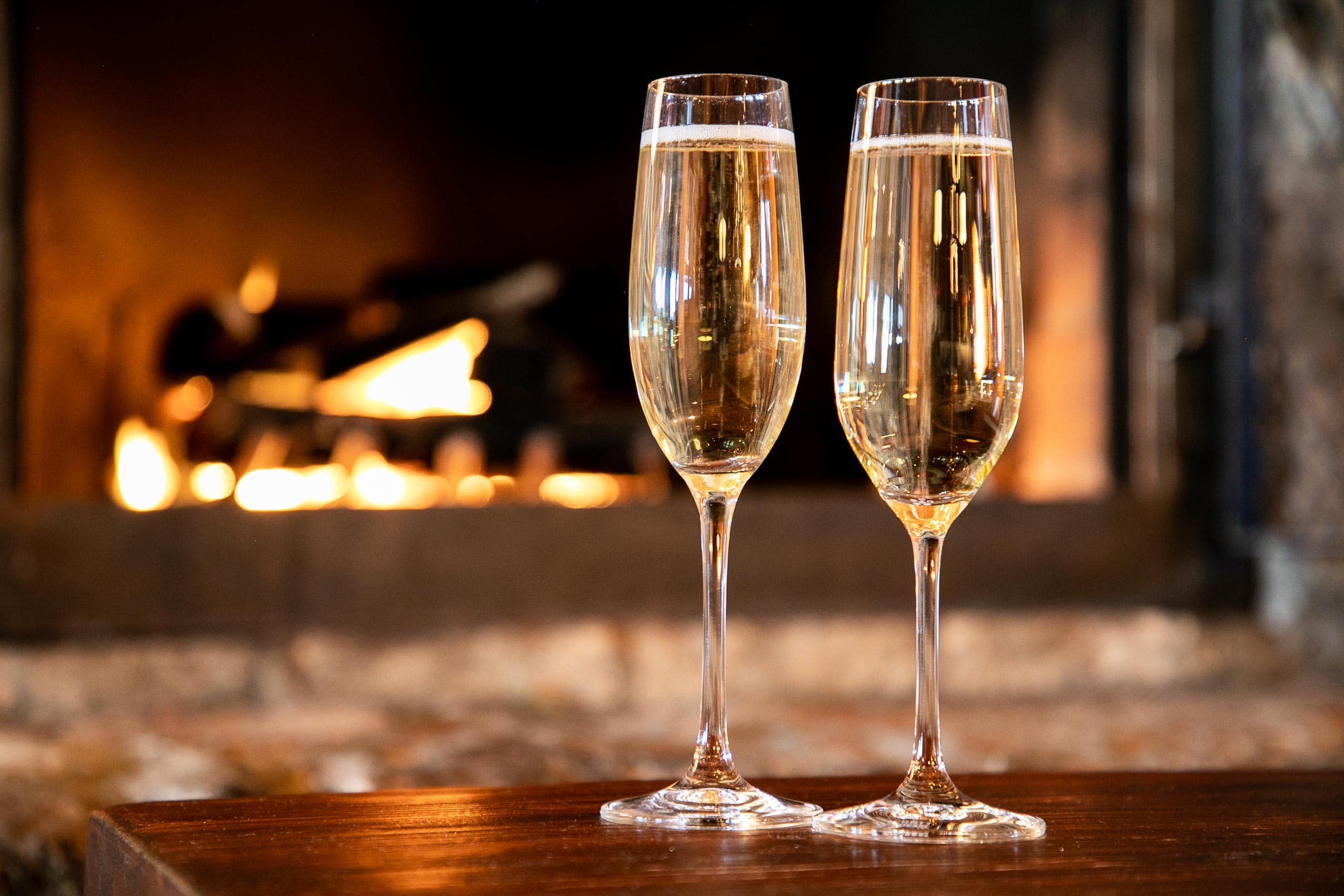 Two champagne glasses on a wooden table with a cozy fireplace in the background.