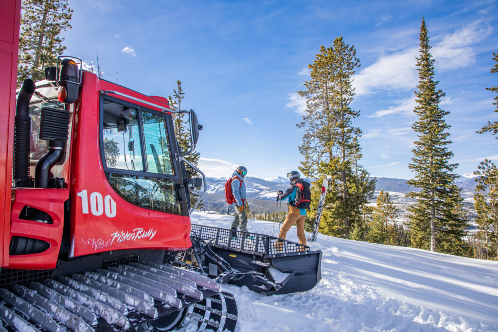 Two skiers stand by a snowcat with their ski gear.