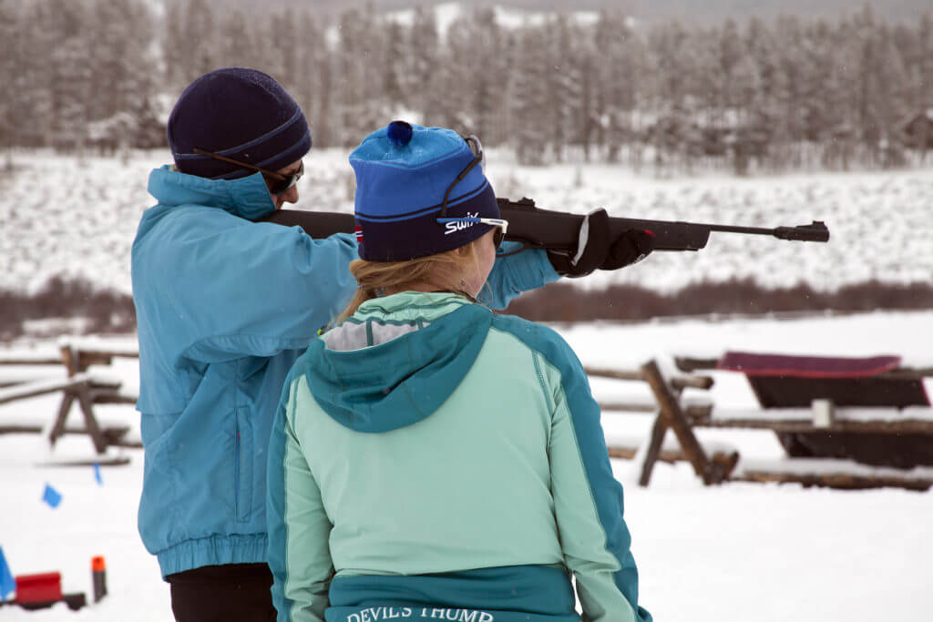 One person holds and aims an air rifle while another person stands beside and watches surrounded by snowy environment